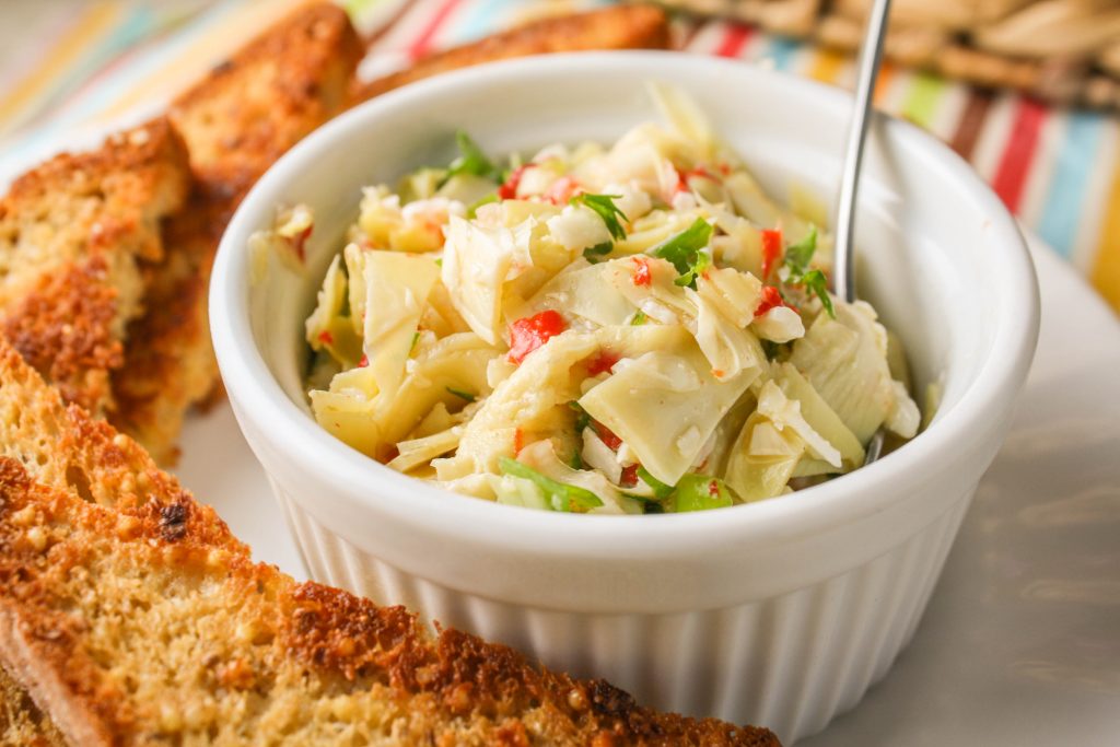 white ramekin filled with chopped artichoke hearts, pimentos, and green onions surrounded by golden brown garlic toasts