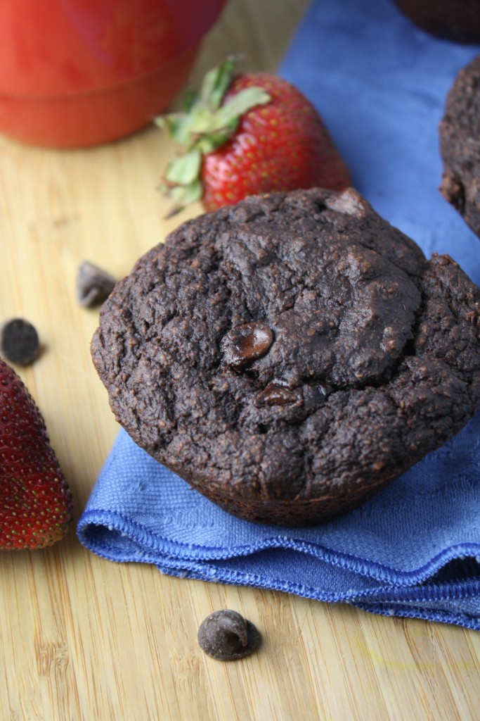  a single rich chocolate muffins studded with chocolate chips and a few chocolate chips sprinkled around a light colored wooden cutting board