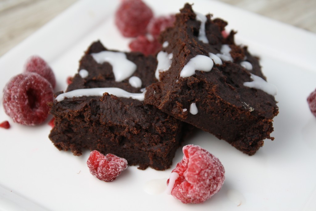 horizontal image of 2 dark chocolate dense brownies with a white coconut butter drizzle and some frozen raspberries scattered on a white plate