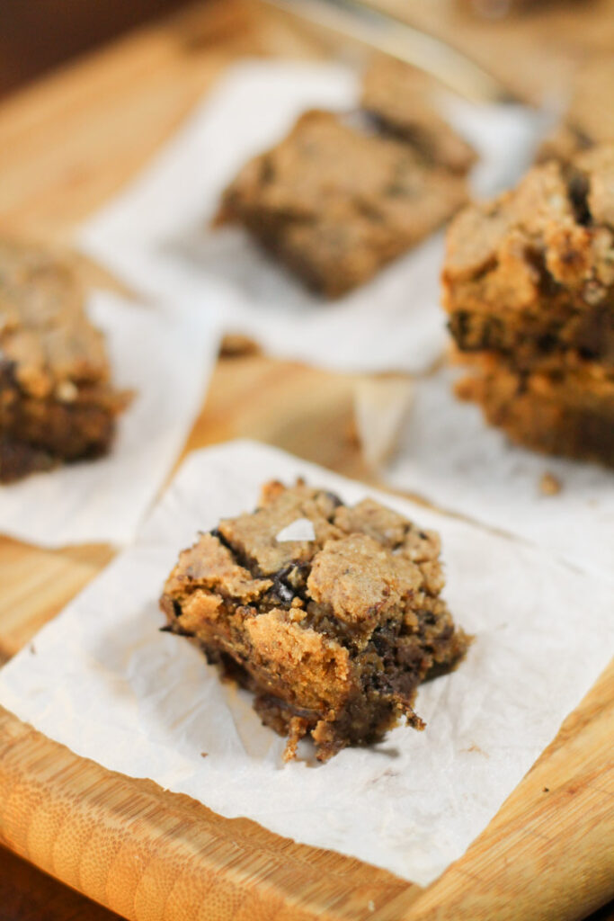 a square of chocolate chip studded blondies on a white piece of parchment paper topped with flaked sea salt