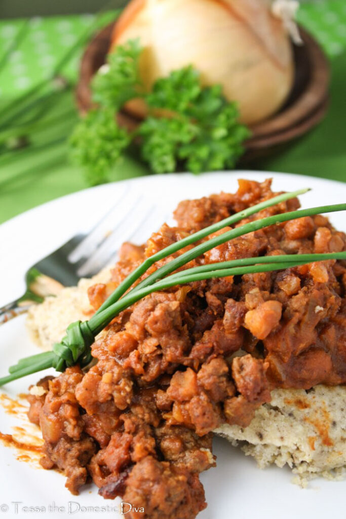 a white plate with a piece of cornbread topped with a bacon, bean, beef, and barbecue sauce skillet meal with a chive garnish