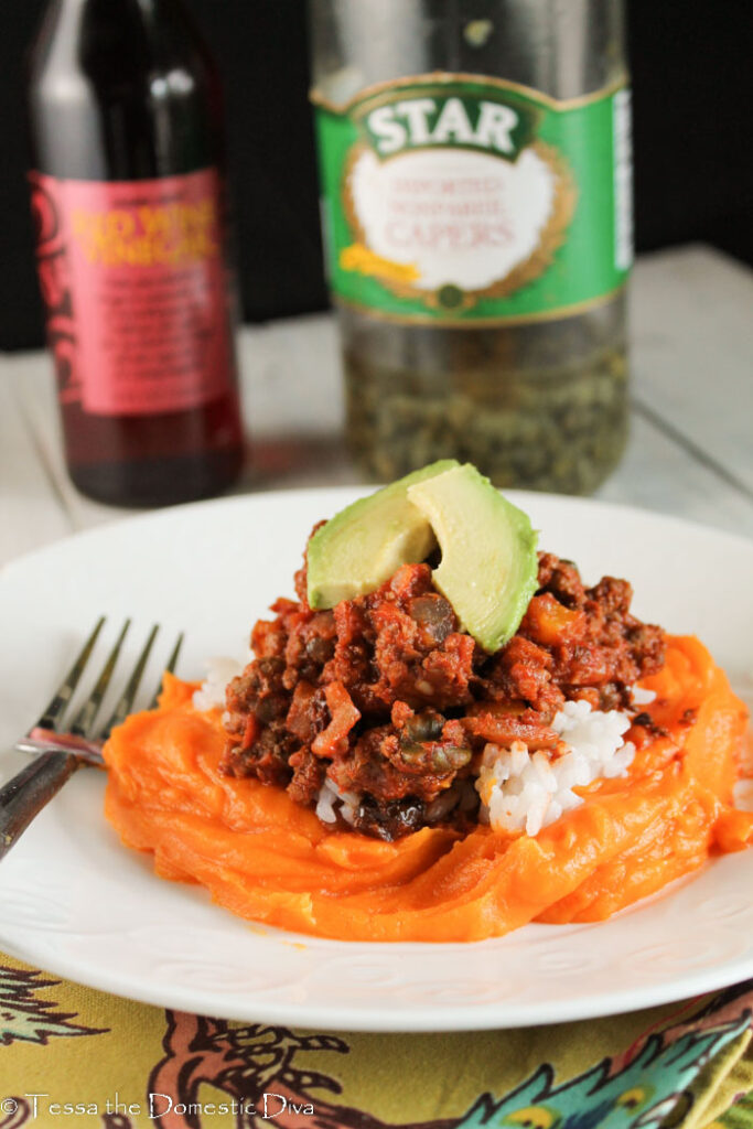 a white plate with Cuban picadillo on a bed of bright orange sweet potato mash with a jar of capers and red wine vinegar in the background