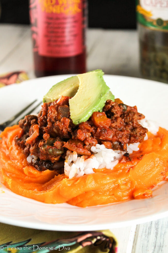 a white plate with garlic mashed sweet potato topped with Cuban picadillo and sliced avocado