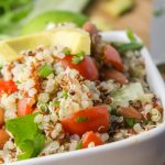 vertical image of fluffy quinoa studded with pinto beans, cilantro, tomatoes, and avocado in a white bowl with lime slices in the bacground