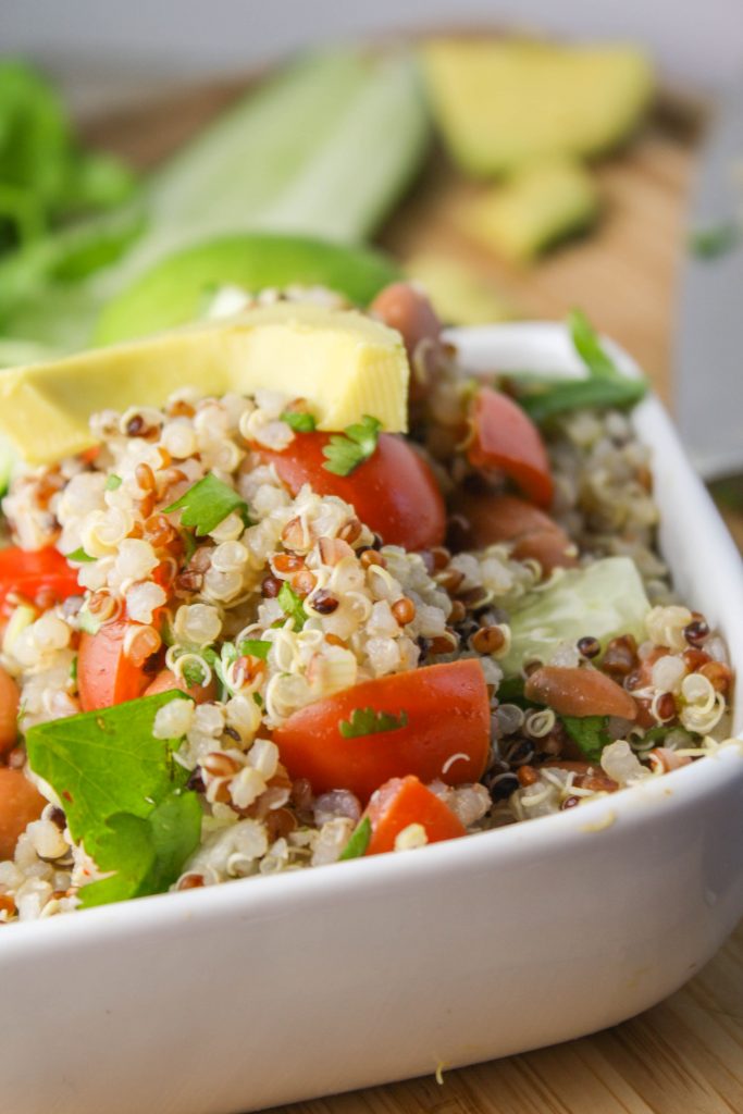 vertical image of fluffy quinoa studded with pinto beans, cilantro, tomatoes, and avocado in a white bowl with lime slices in the bacground