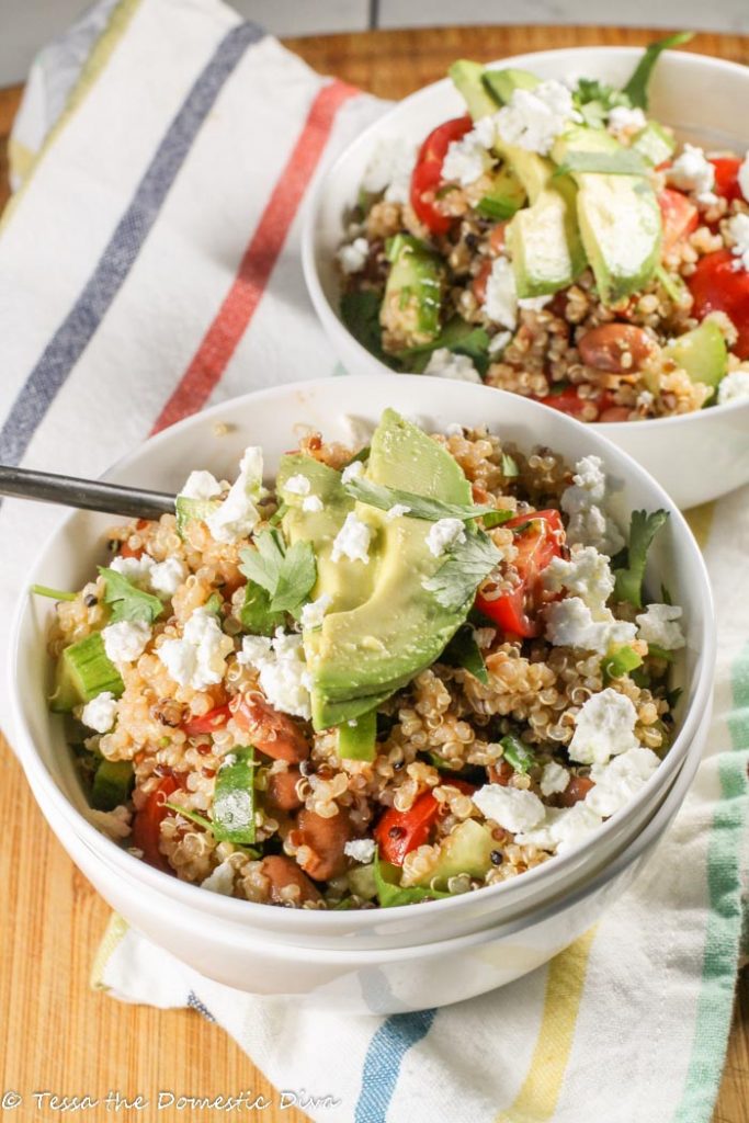 two white bowls filled with quinoa salad garnished with avocado, feta, and cilantro