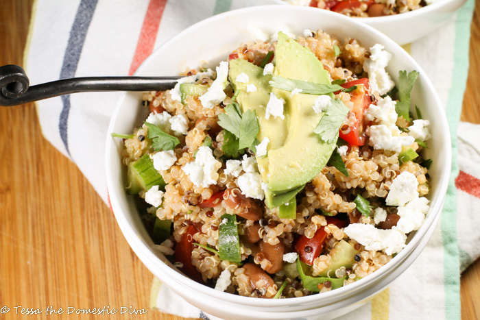 a single white bowl atop a bamboo cutting board filled with quinoa, tomatoes, beans, and cucumber topped with feta, avocado, and cilantro