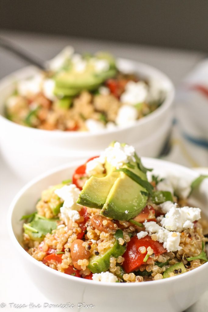 two white bowls filled with cooked quinoa, tomatoe, cucumber, and pinto beans and garnished with avocado, cilantro, and feta