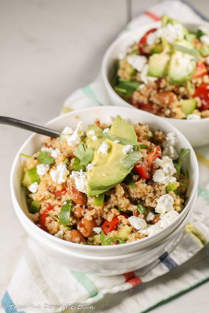 white bowls filled with cooked quinoa, tomatoes, beans, and cucumbers garnished with cilantro, feta and some sliced avocado