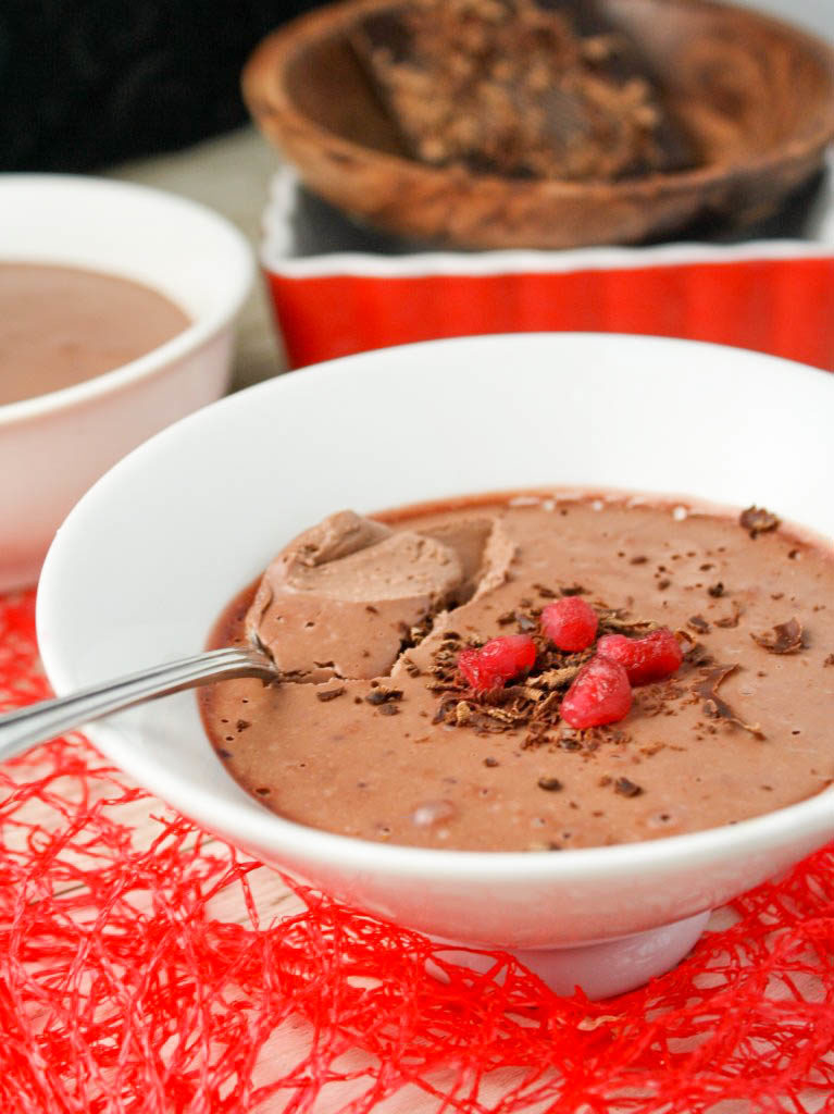 white bowl with a tapered bottom filled with a pale brown thick pudding garnished with shaved chocolate and 5 pomegranate seeds