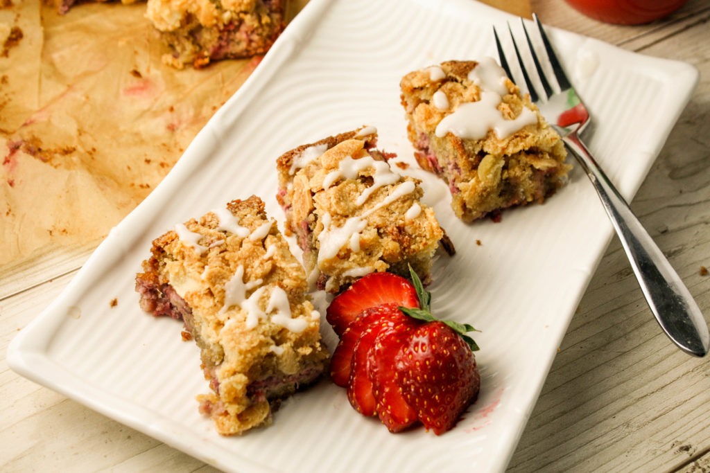 three rectangular slices of a strawberry studded paleo coffee cake with a coconut butter drizzle and fresh cut berries on a white plate