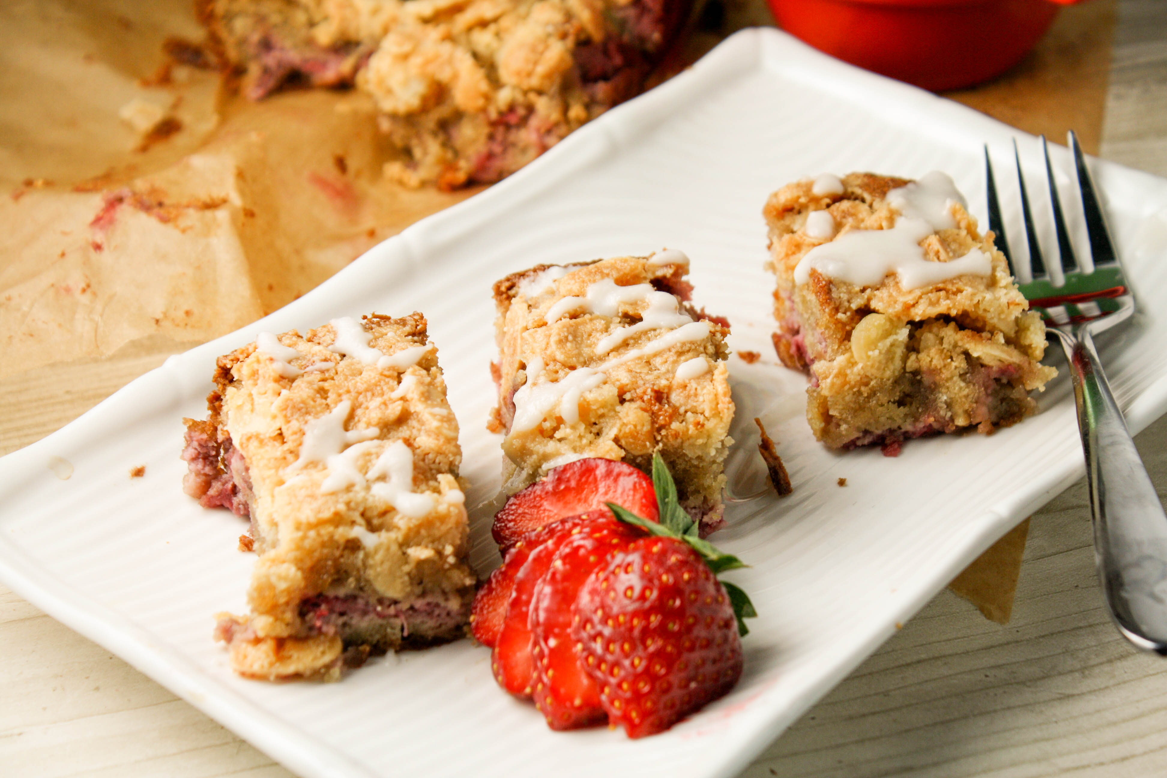 three rectangle shaped slices or a coffee cake studded with diced starwberries on a white plate with a fresh starwberry garnish and white drizzle