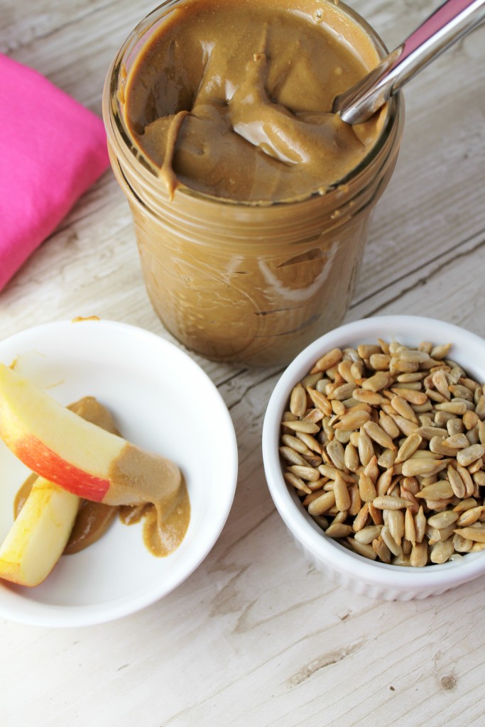 fresh homemade creamy sunflower seed butter in a mason jar with a white bowl of whole sunflower seeds and a sliced apples with a red skin