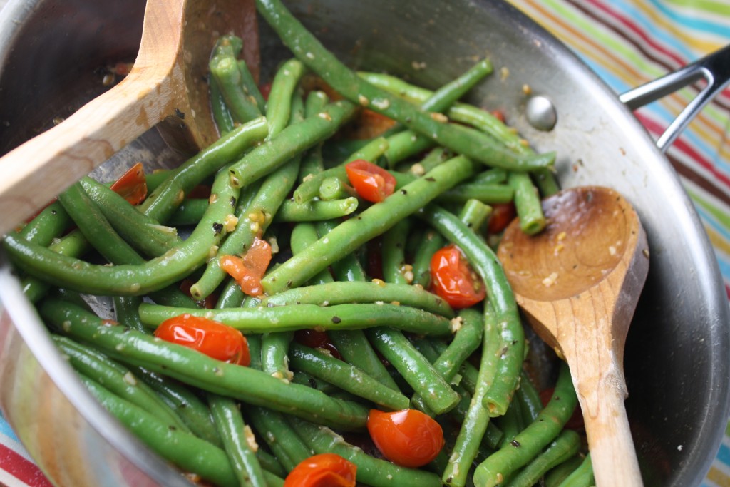 overhead shot of a stainless steel pan filled with steamed green beans, tomatoes, and fresh basil and two wooden serving spoons