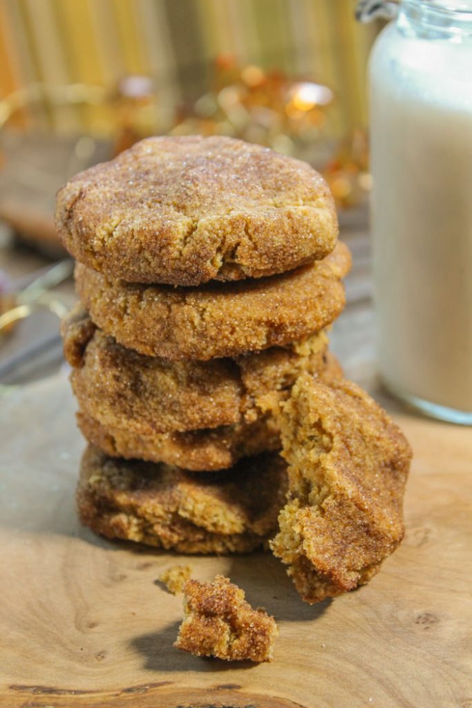 orange hued stack of pumpkin cookies coated in cinnamon and sugar atop a natural wooden board and a vintage milk glass in background