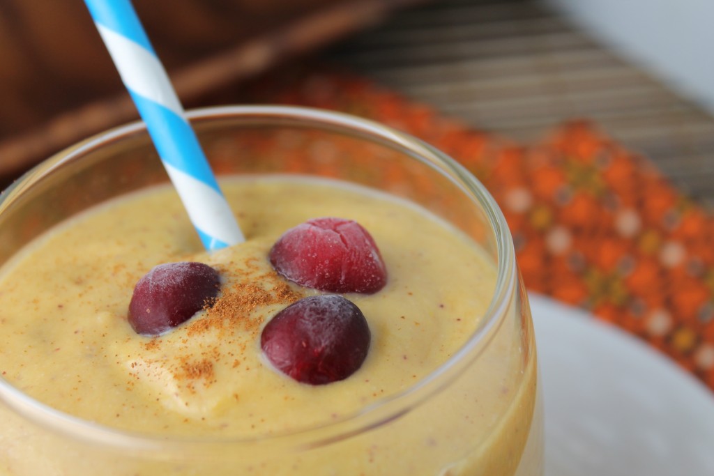horizonatal image close up of a creamy orange hued pumpkin smoothie topped with a sprinkle of cinnamon and fresh whole cranberries with a pumpkin orange floral cloth