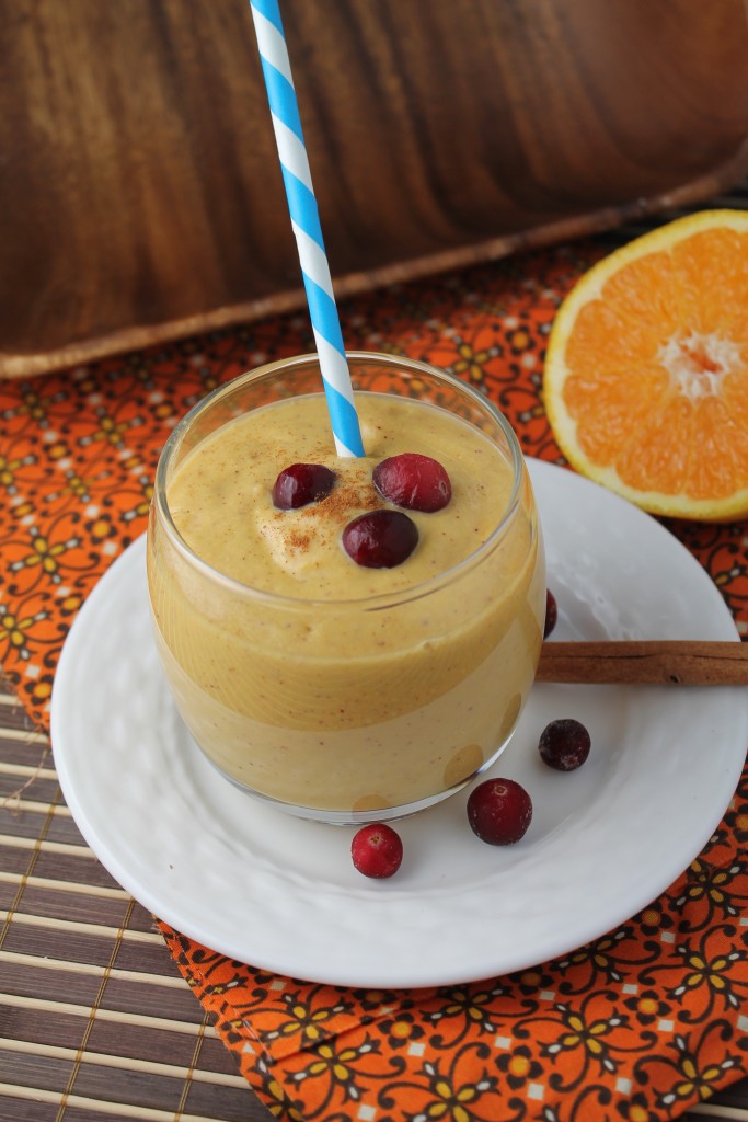 a clear glass of a pumpkin hued thick smoothie with orange, cinnamon, fresh cranberries, and pumpkin on a white plate with an orange cloth