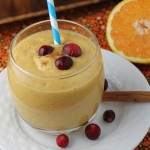 a creamy orage hued pumpkin smoothie topped with fresh cranberrie and sliced orange half in background