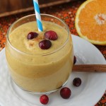 a creamy orage hued pumpkin smoothie topped with fresh cranberrie and sliced orange half in background