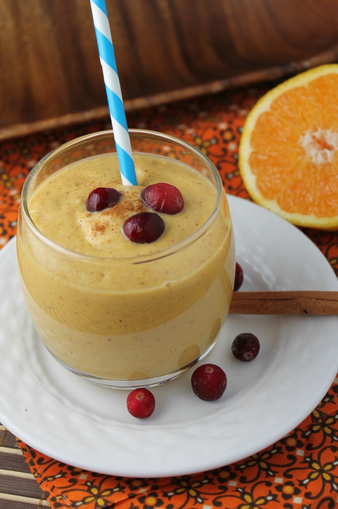 a creamy orange hued pumpkin smoothie topped with fresh cranberries and sliced orange half in background