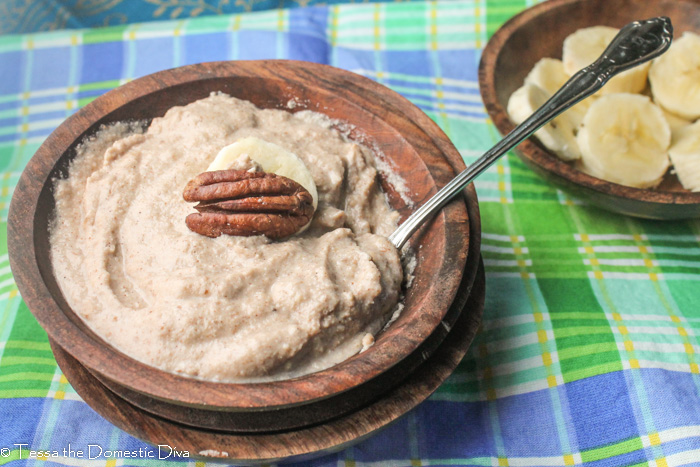a dark wooden bowl filled with a nut porridge topped with a pecan half and a bowl of sliced fresh banana