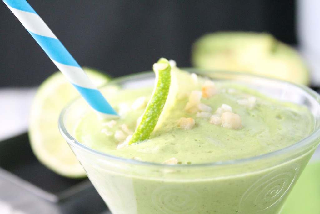 whole30 lime smoothie close up at eye level in a clear round glass with a blue and white paper straw, a garnish of sliced lime and chopped macademia nuts