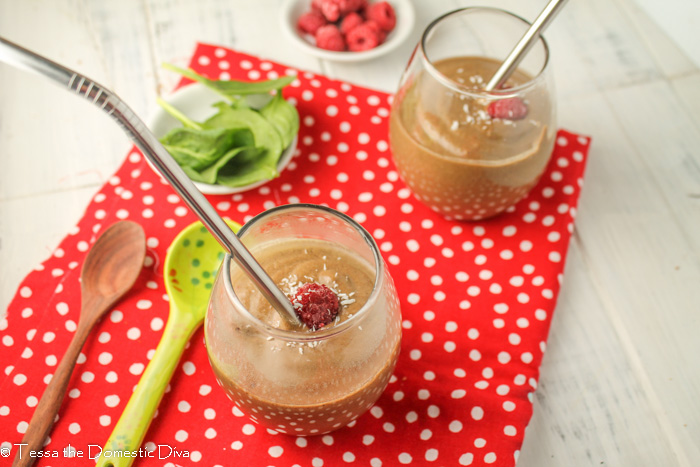 birds eye view of two clear glasses with a thick chocolate smoothie topped with a raspberry and next to fresh spinach on a red cloth