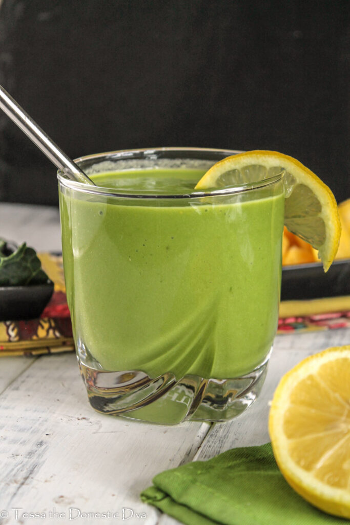 a clear glass with a stainless straw filled with a vibrant green smoothie with a slice of lemon on the glasses edge at eye level