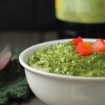 eye level white bowl filled with a vibrant green pesto sauce with olive oil, kale in background