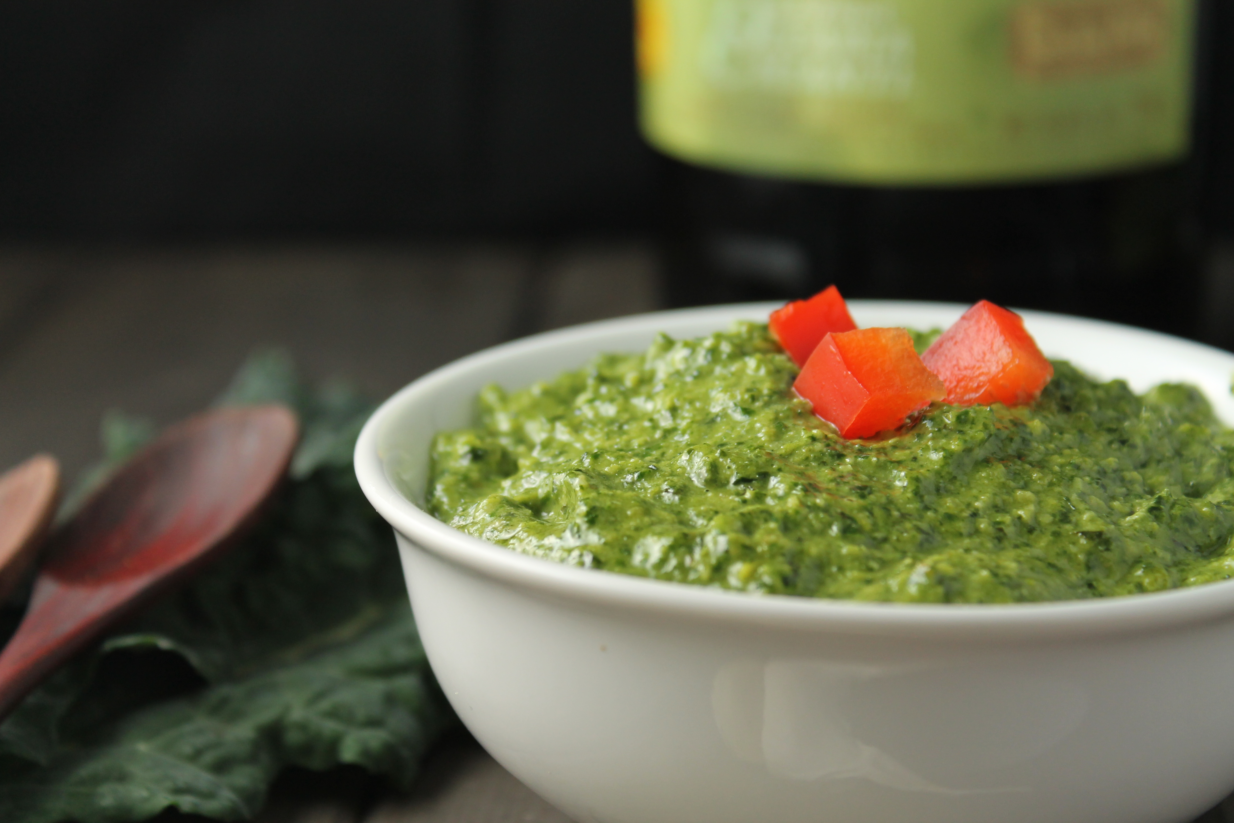 eye level white bowl filled with a vibrant green pesto sauce with olive oil, kale in background