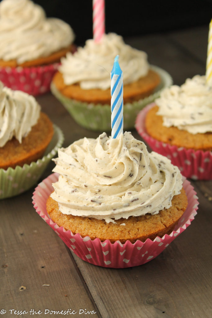 vanilla cupcakes arranged on a dark wooden surface with swirled vanilla frosting and a single birthday candle in each cupcake