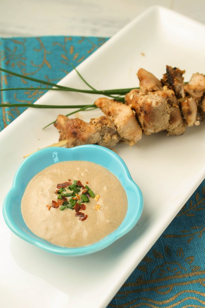 a white plate with a blue bowl of peanut sauce and chicken satay.