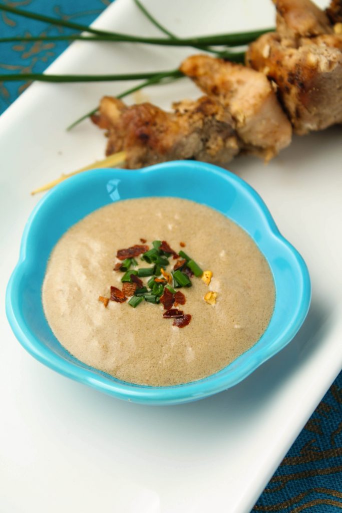  peanut sauce in a turquoise dipping dish with chicken skewers and a chive and red pepper flake garnish.