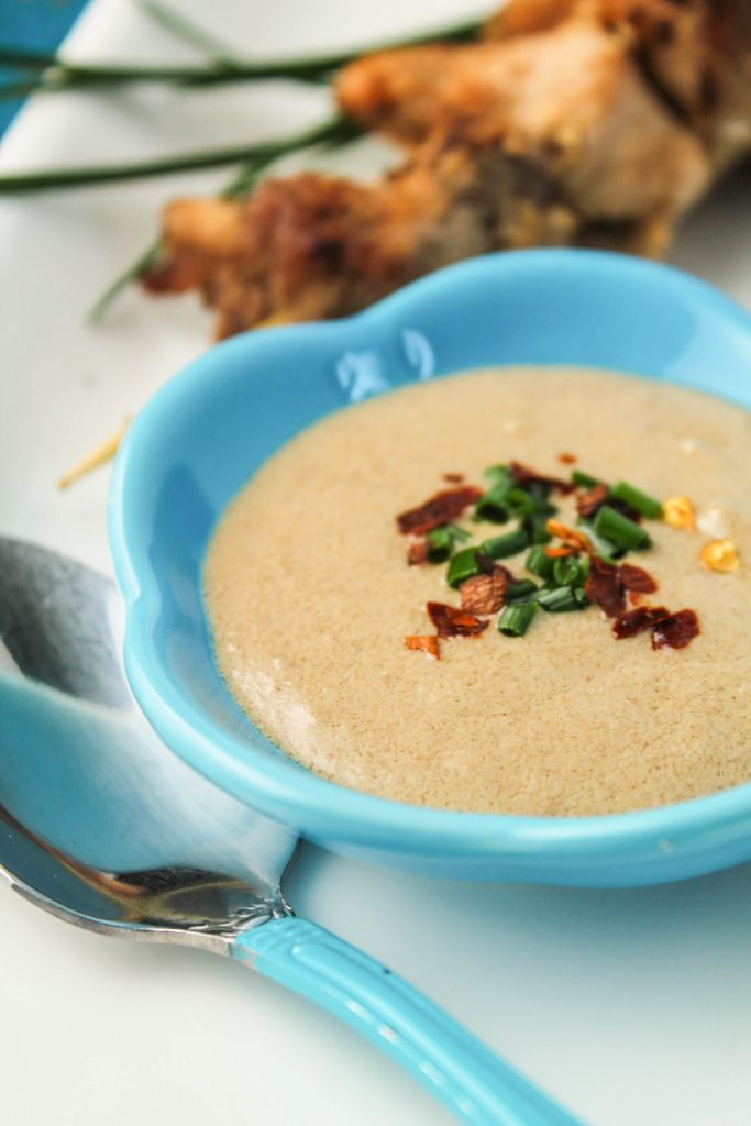 close up of a creamy peanut sauce in a small turquoise dipping bowl with a garnish of chives and red pepper flakes.