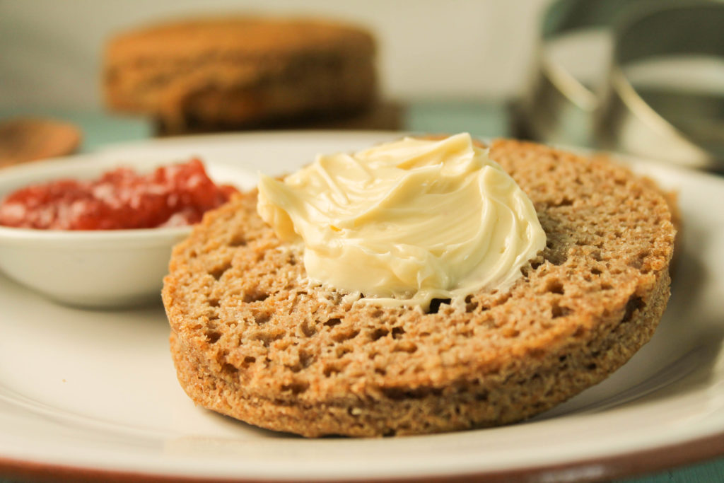 horizontal image of a gluten free teff bread english muffin slice slathered in butter with a bowl of jelly in background