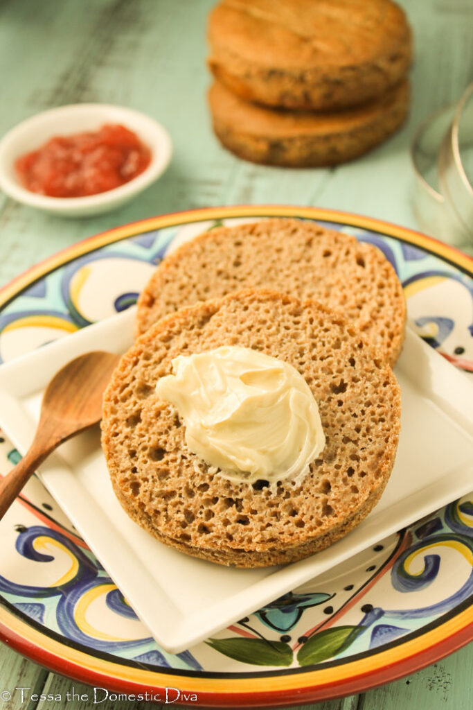 front view of a festive plate and a sliced teff English muffin atop a square white plate with a pat a butter