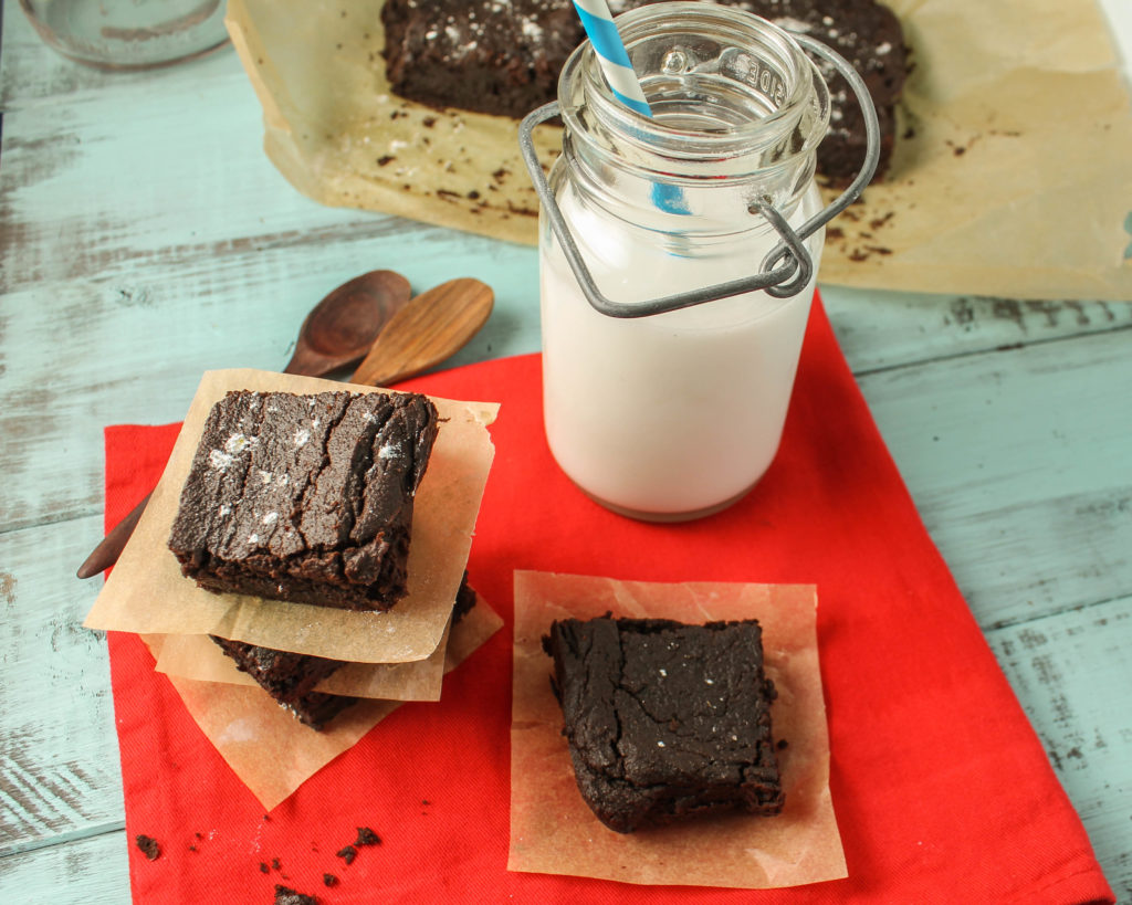 overhead shot of paleo nut free egg free brownies on a red cloth and blue wooden board with two wooden spoons and a glass of coconut milk.