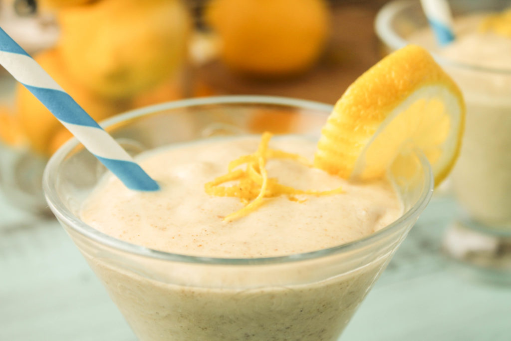 a keto creamy lemon smoothie in a clear glass topped with lemon zest, a lemon slice, and blue and white paper straw