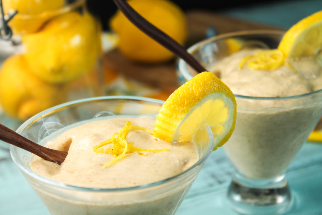 2 clear glasses of a pale yellow lemon smoothie garnished with lemon zest, a lemon slice, and using a blue an white paper straw