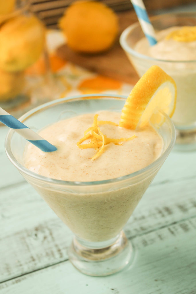 two clear glasses filled with a plae yellow creamy smoothie with fresh lemon zest and lemon slice on the glass edge