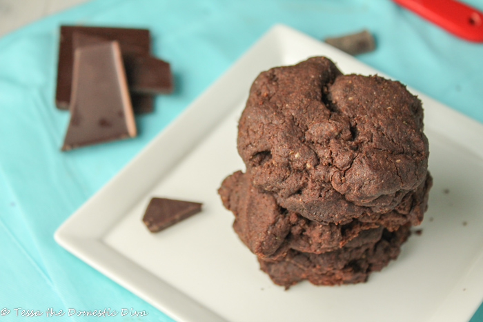 three stacked double chocolate chip cookies on a white late with a red clay measuring spoon in the background