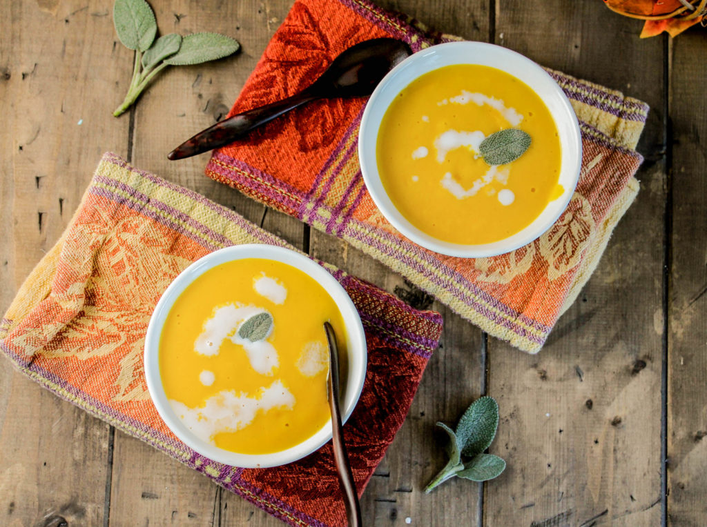 two white bowls of orange hued creamy pumpkin soup from over head with a coconut cream swirl and sage leaf on a dark wood surface