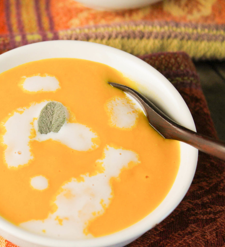 close up of a creamy pumpkin soup in a white bowl with a dark wooden spoon on fall themed napkins and a sage leaf and coconut cream garnish