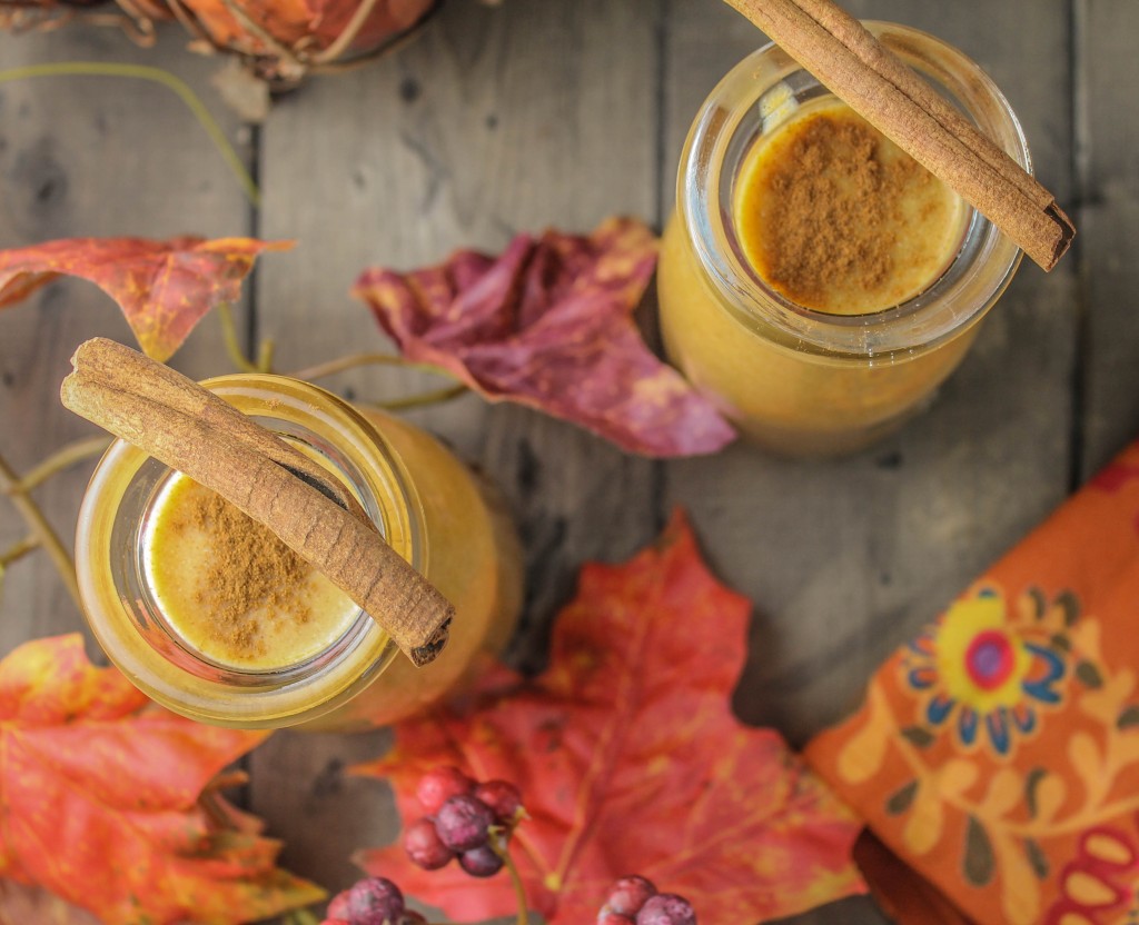 from overhead looking down on two clear glass jars filled with an orange pumpkin smoothie and a cinnamon stick resting on the glass's edge on a dark wooden surface 