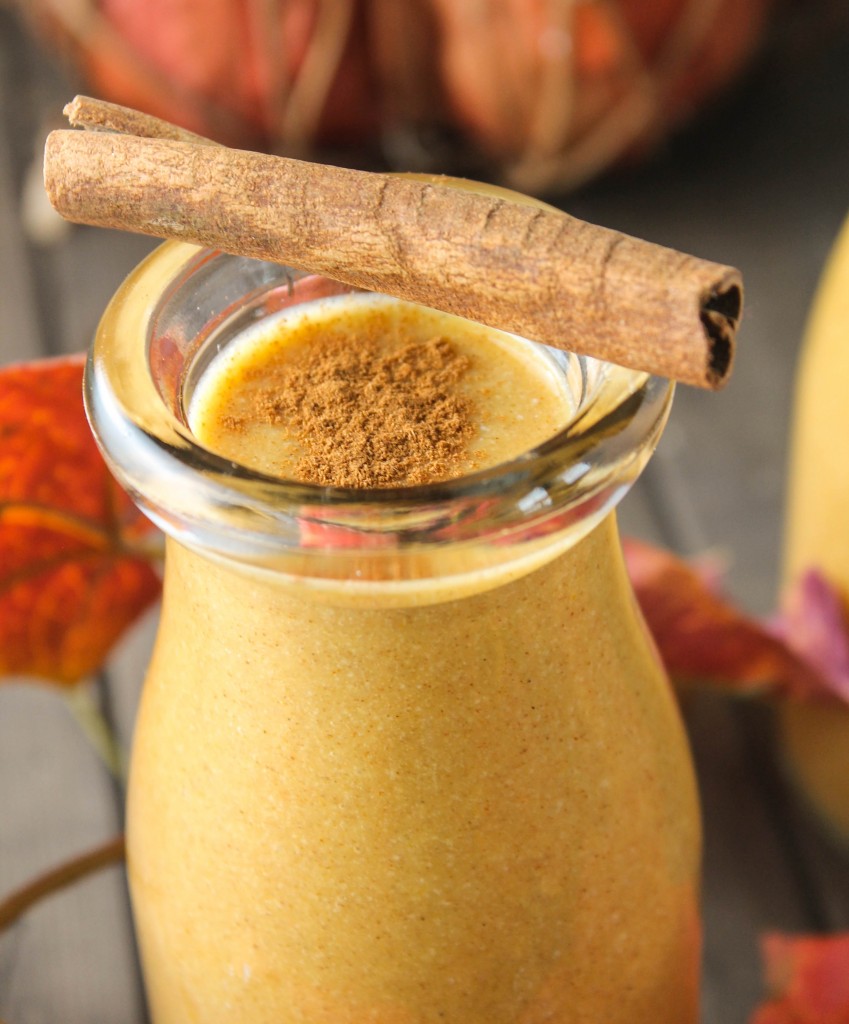 a close up of a clear glass filled with an orange colored pumpkin smoothie topped with a sprinkle to cinnamon and a cinnamon stick resting on the glass's edge