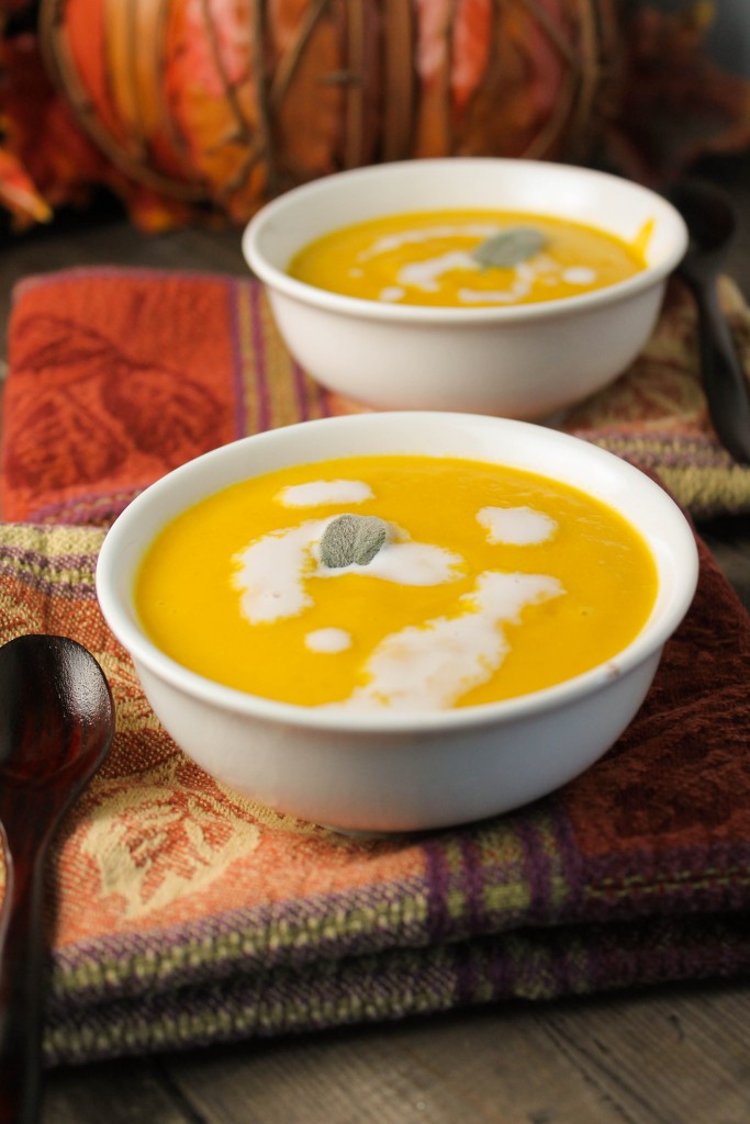 two white bowls on top of fall colored napkins filled with orange pumpkin soup and dollops of creams and a garnish of sage