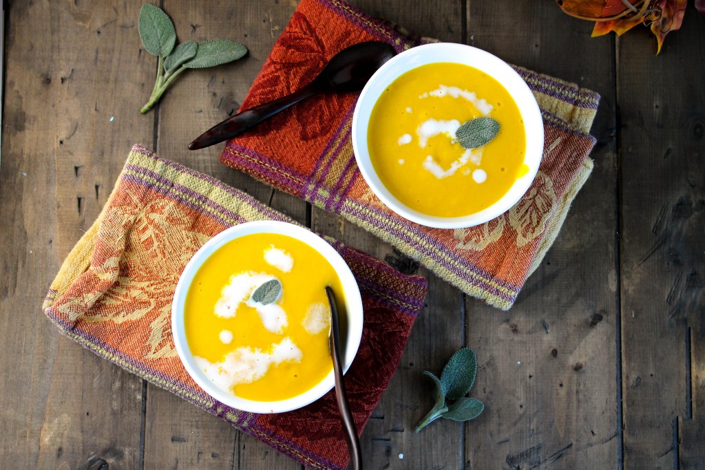 birdseye view of two white bowls filled with pumpkin soup and swirled with white cream and a fresh sage leaf