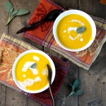 birdseye view of two white bowls filled with pumpkin soup and swirled with white cream and a fresh sage leaf