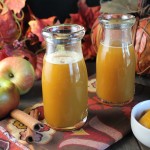 two vintage milk glasses filled with an orange hued apple cider drink on a dark wooden surface with fresh apples and fall leaves