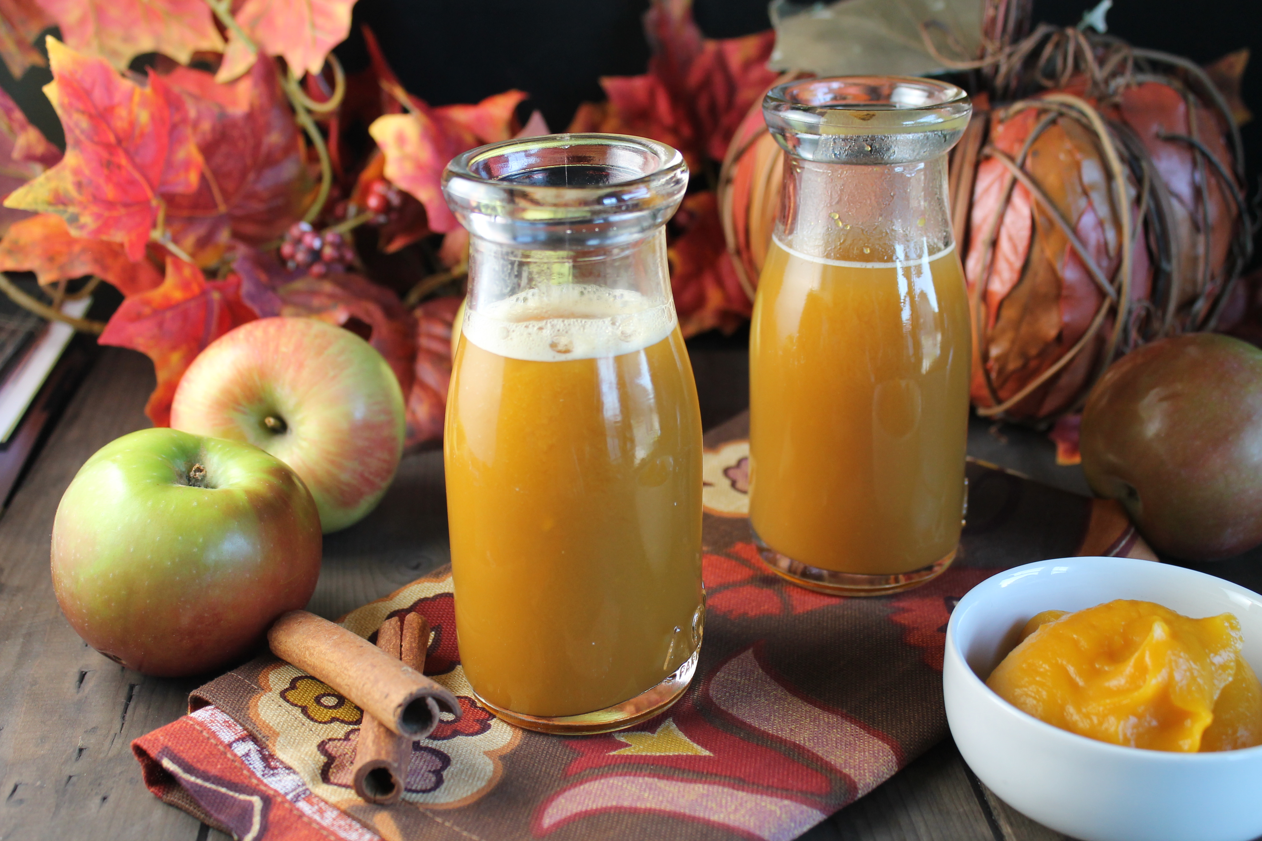 two vintage milk glasses filled with an orange hued apple cider drink on a dark wooden surface with fresh apples and fall leaves